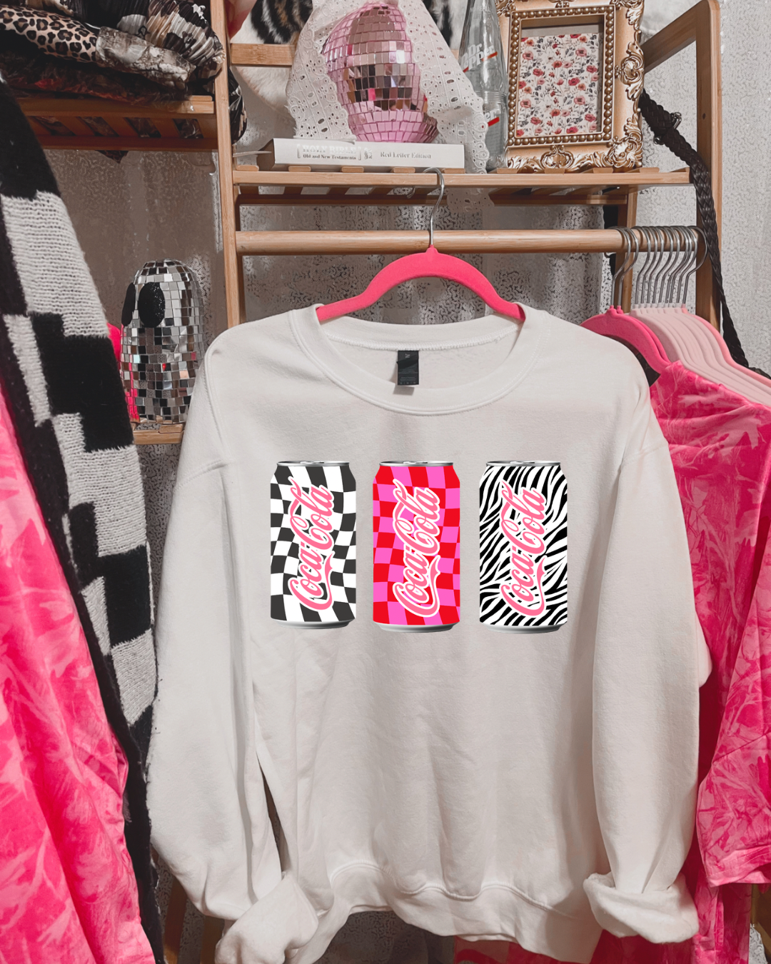 White sweatshirt with colorful 'Coca-Cola' graphics on a hanger against a clothing store background.